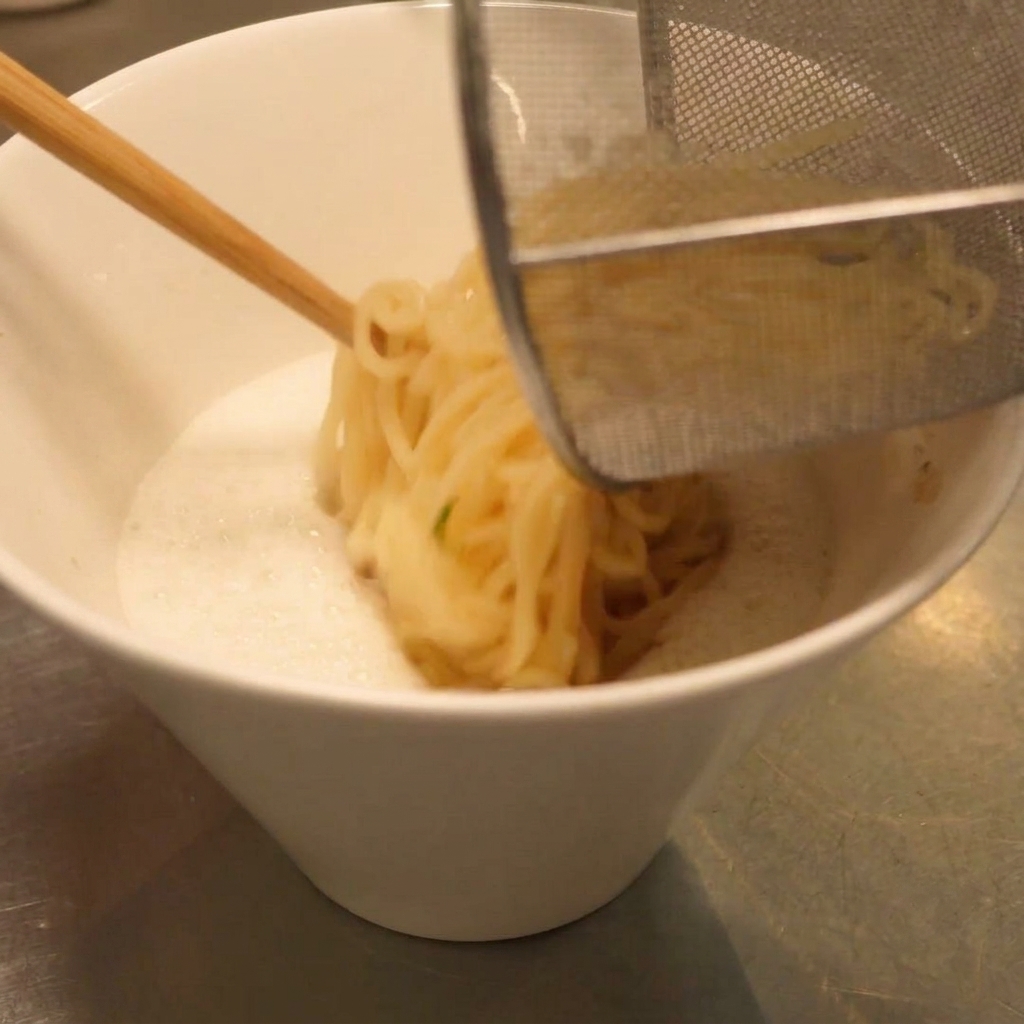 Odcedzony makaron ramen przekładany do miski z kremową zupą.