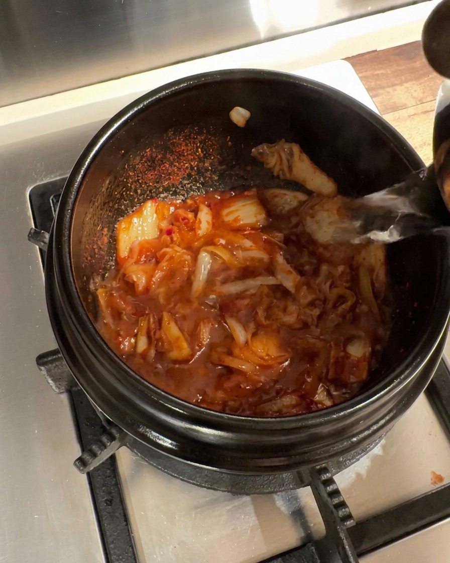 Wrząca woda wlewana do kamiennego garnka z doprawioną mieszanką kimchi i cebuli.
