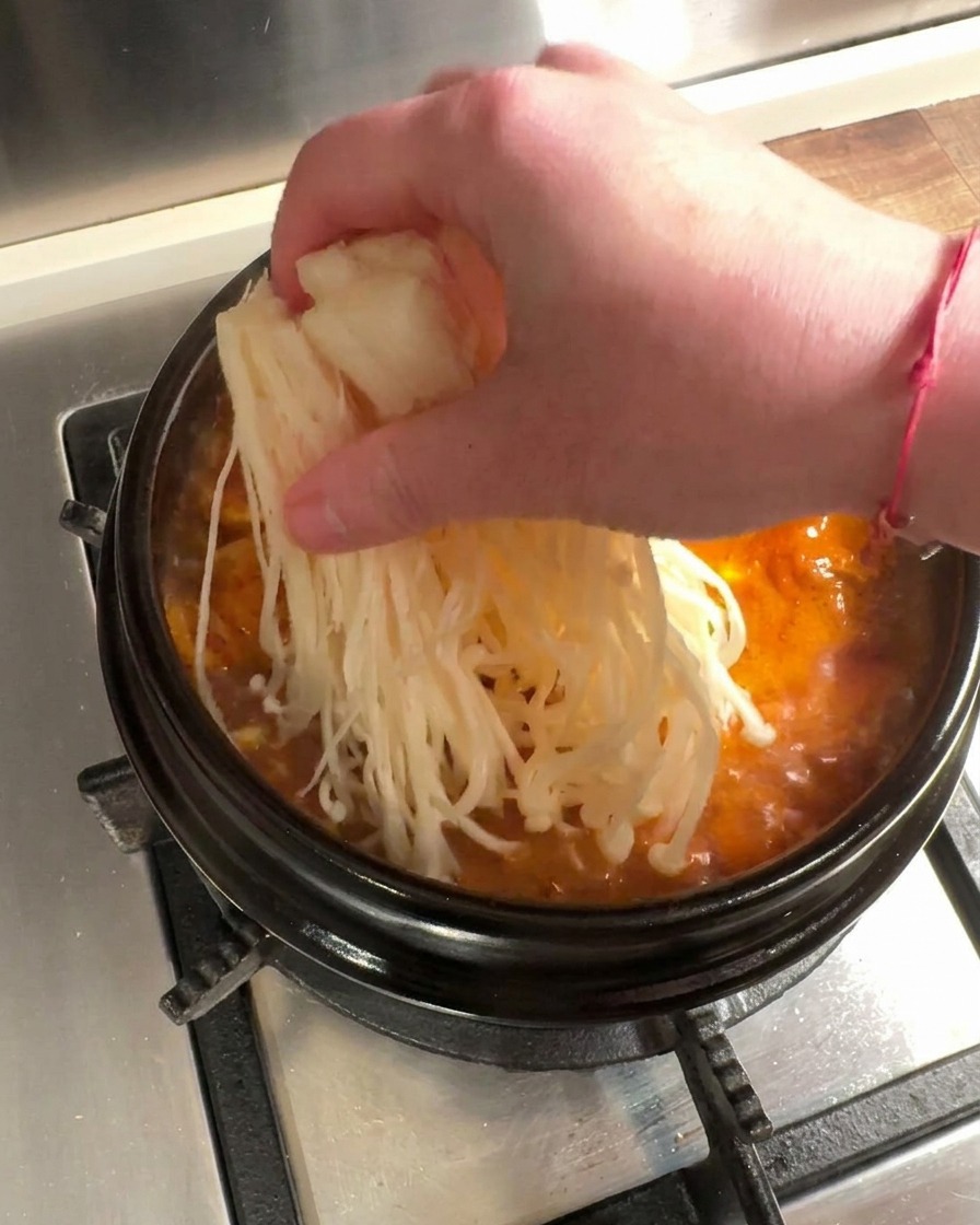 Świeże grzyby enoki i cienko pokrojona surowa wołowina umieszczane w bulgoczącej czerwonej zupie kimchi.