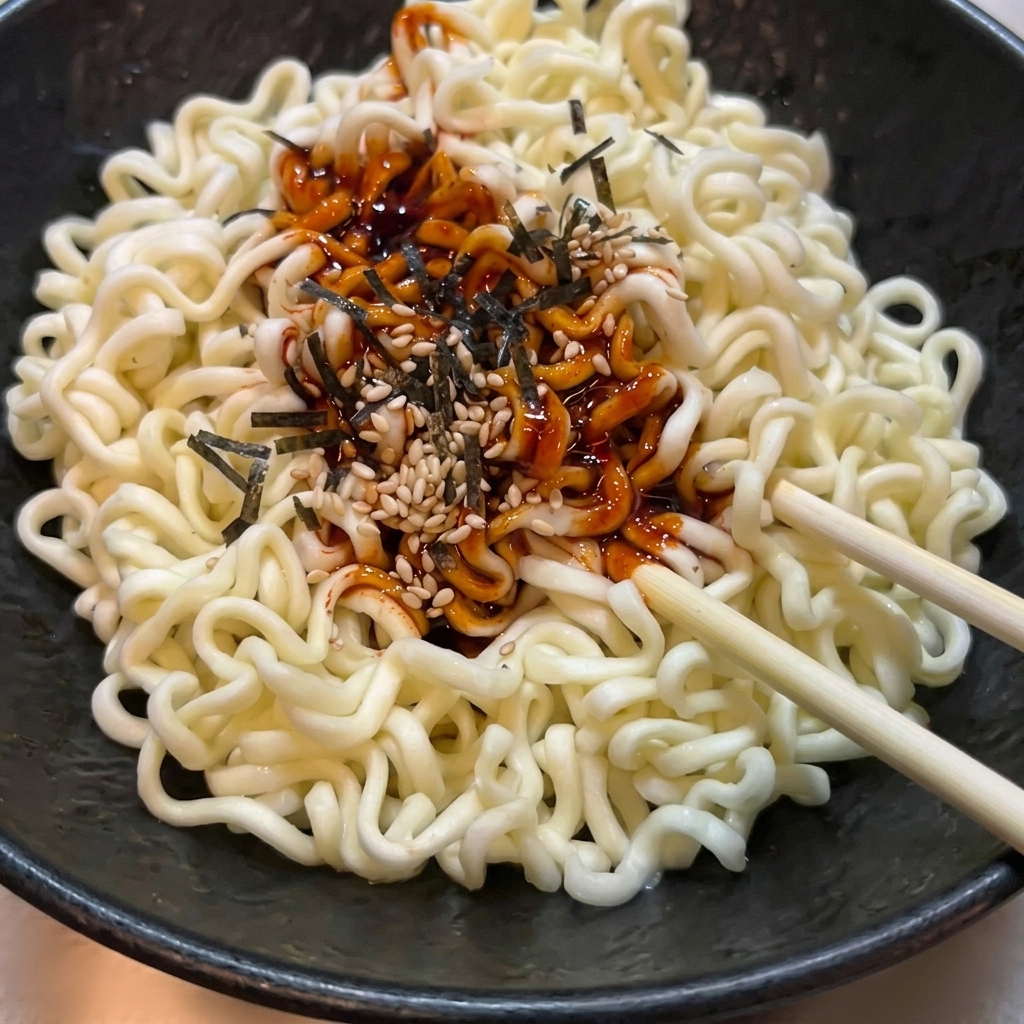 Ugotowany makaron ramen w czarnej misce mieszany z czerwonym pikantnym sosem, sezamem i wodorostami.