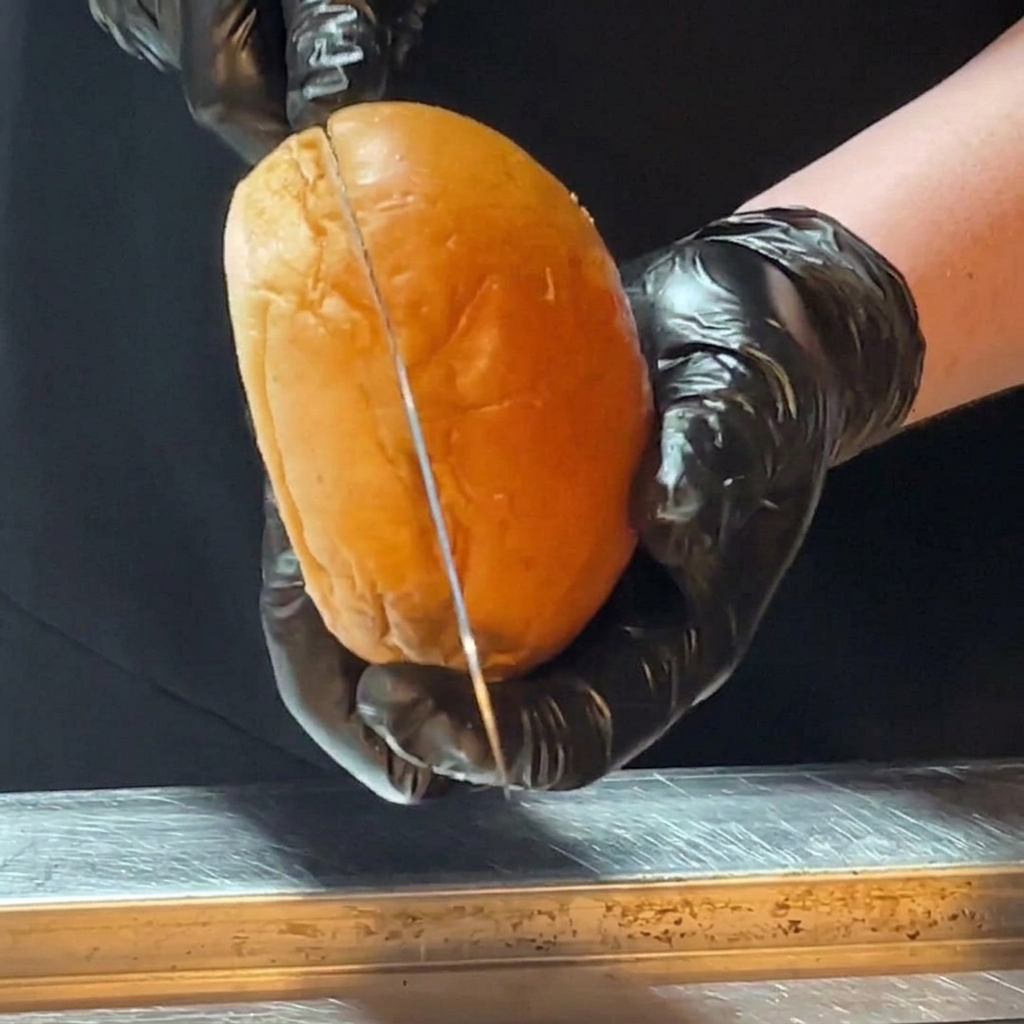 Dłonie w rękawiczkach przekrawają bułkę brioche długim nożem.