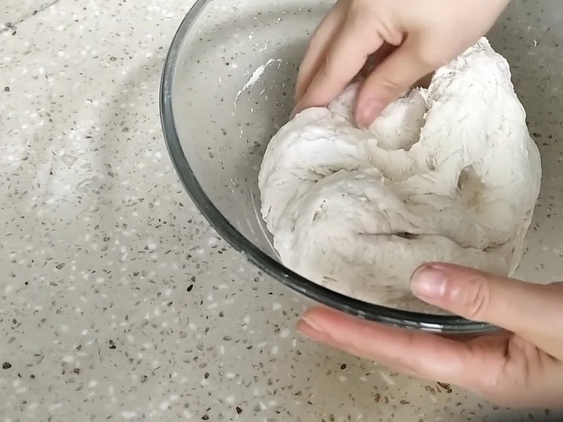 Ręce wyrabiające grudkowatą kulę ciasta w szklanej misce.