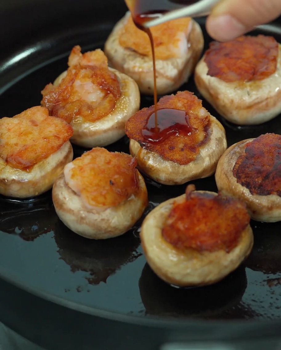 Wlewanie mieszanki ciemnego sosu sojowego i soku z pieczarek na faszerowane pieczarki na patelni.
