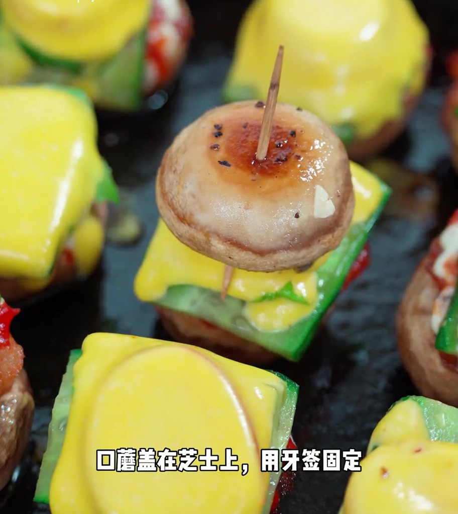 Gotowy mini burger grzybowy przebity wykałaczką, pokazujący ułożone warstwy pieczarki, ogórka, sałaty, pomidora, sera i drugiego kapelusza pieczarki.