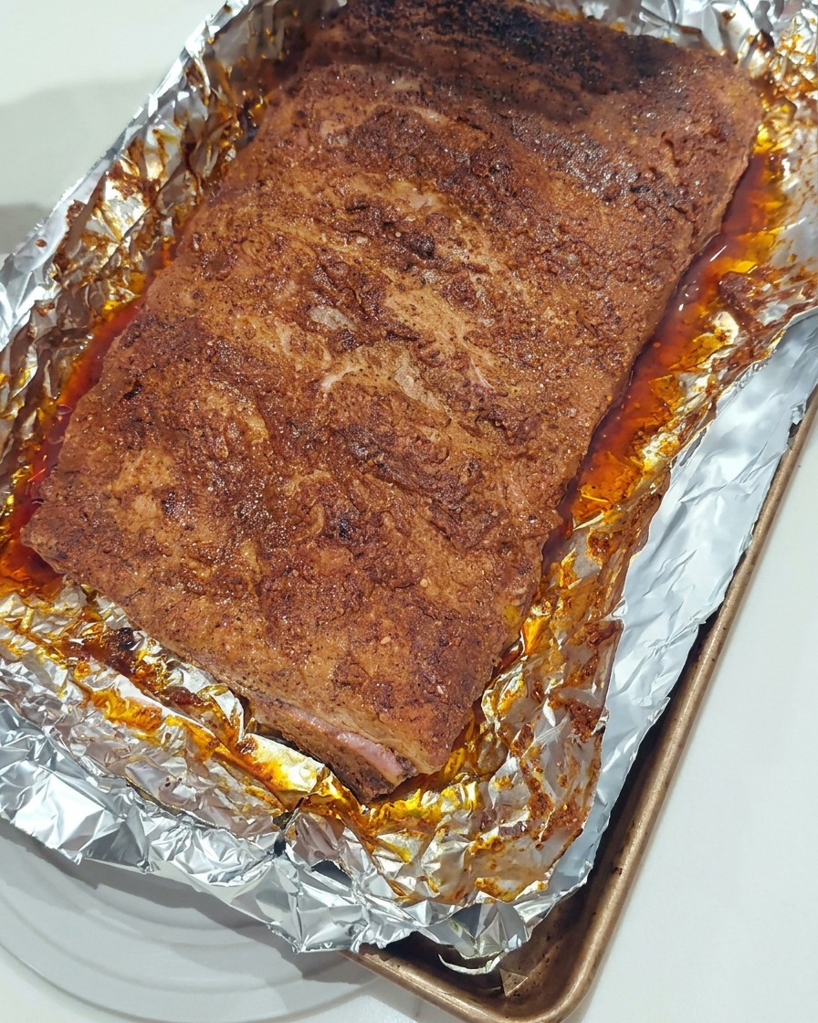 Silikonowy pędzel nakładający lśniącą warstwę gęstego sosu BBQ na powierzchnię upieczonych żeberek w blaszce wyłożonej folią.