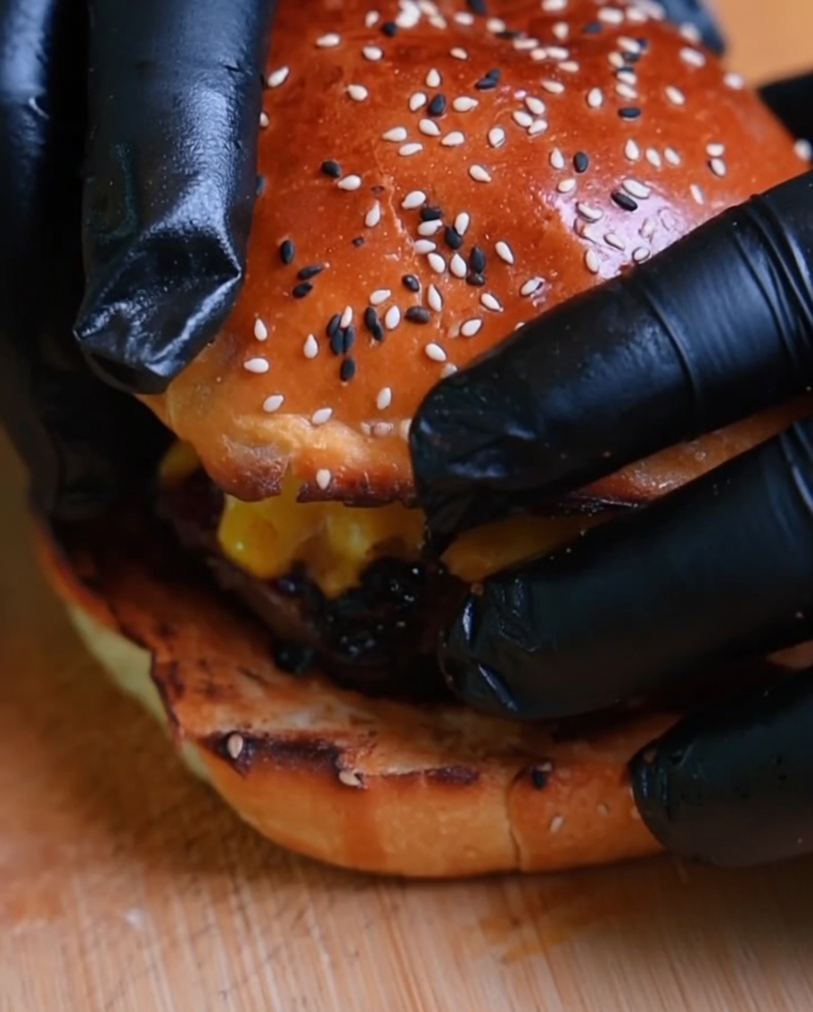 Zbliżenie na burgera, na którym kładziona jest górna część bułki przez ręce w rękawiczkach.