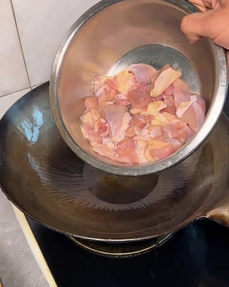 Surowe kawałki udek kurczaka wrzucane z metalowej miski na gorący, naoliwiony wok.