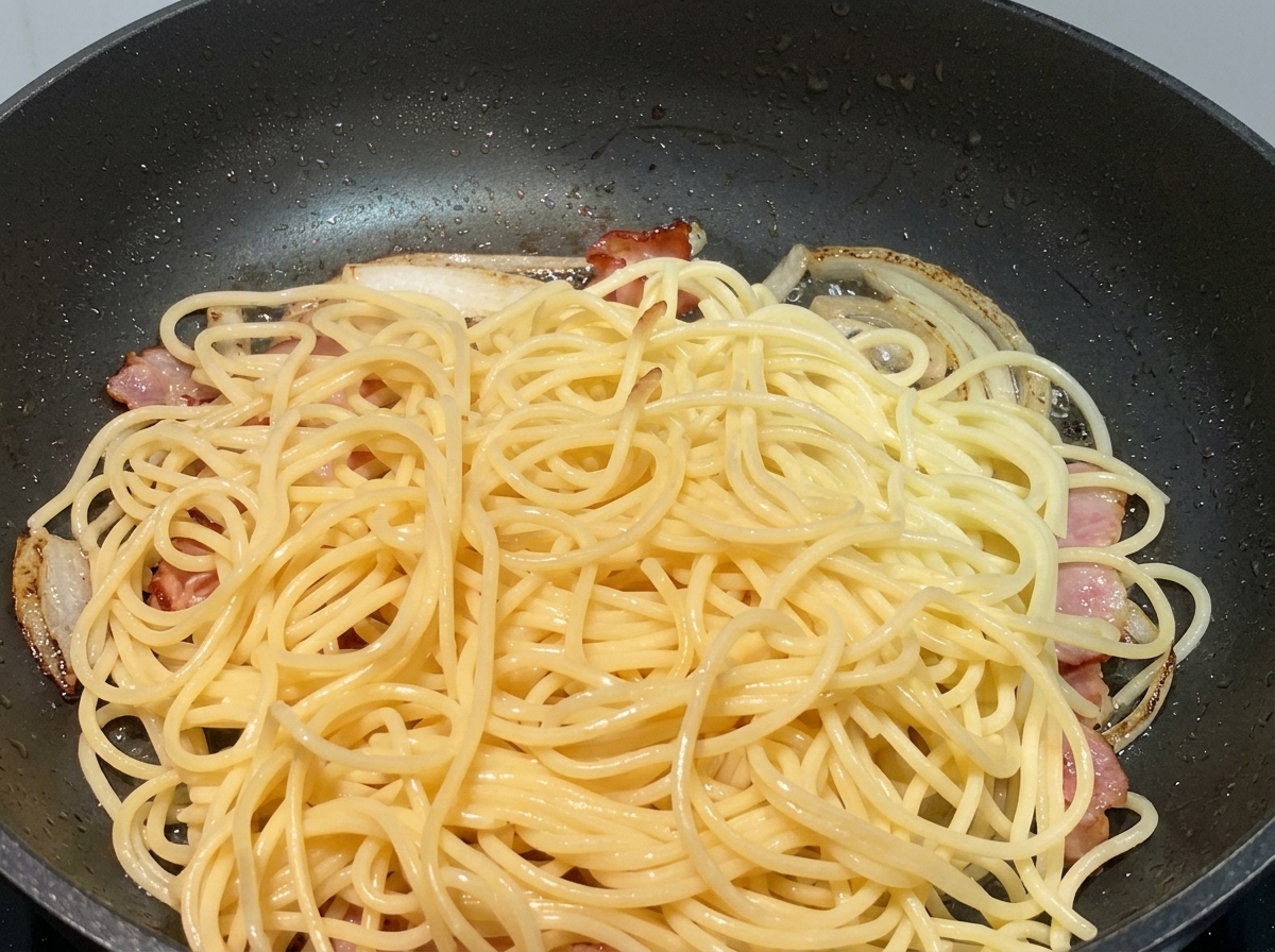 Ugotowany makaron spaghetti na podsmażonej cebuli i kawałkach boczku na czarnej patelni.
