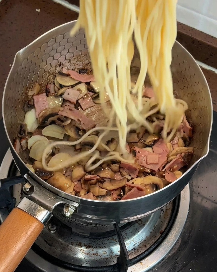 Ugotowane spaghetti dodawane na patelnię z mieszanką podsmażonych grzybów i boczku.