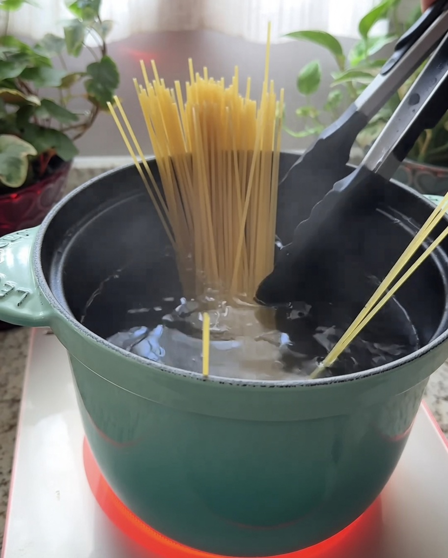 Suchy makaron spaghetti zanurzany w turkusowym garnku z wrzącą wodą.