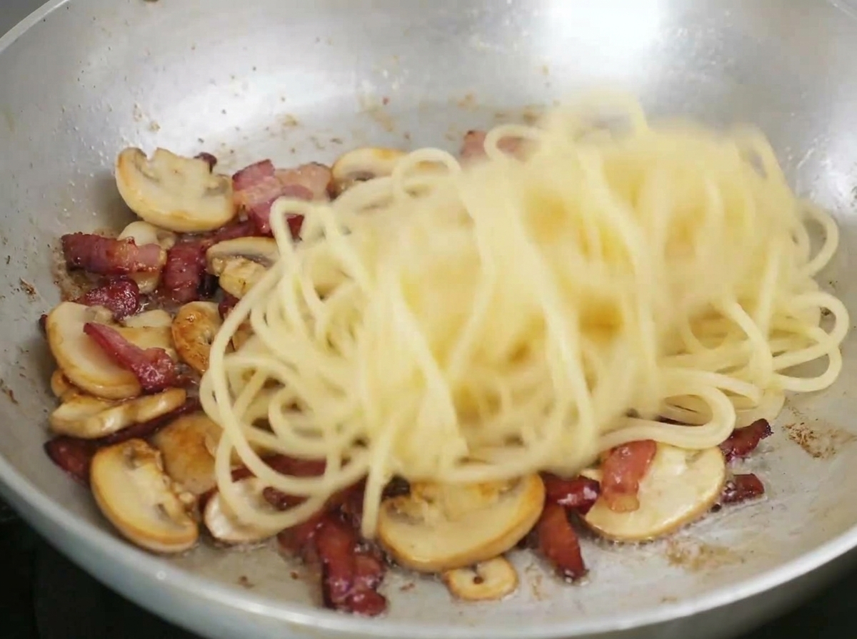 Spaghetti mieszane na patelni z usmażonym boczkiem i pokrojonymi grzybami.