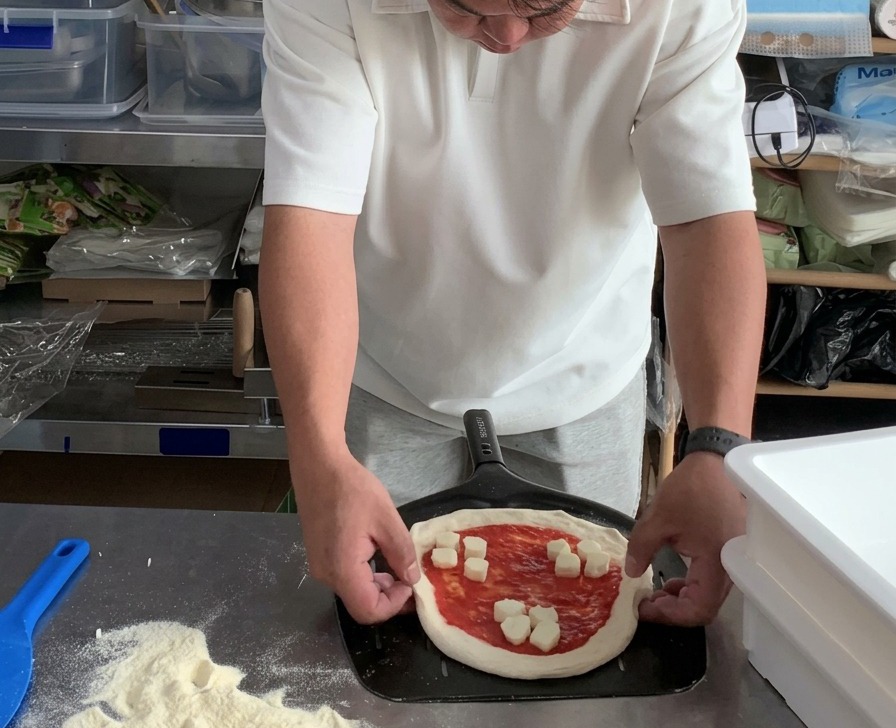 Dłonie poprawiające brzegi surowej pizzy spoczywającej na metalowej łopacie do pizzy.