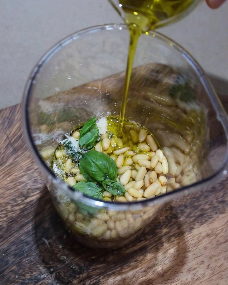 Zbliżenie na składniki pesto, w tym świeżą bazylię, orzeszki piniowe i parmezan, blendowane w naczyniu z wąskim strumieniem wlewanej oliwy.