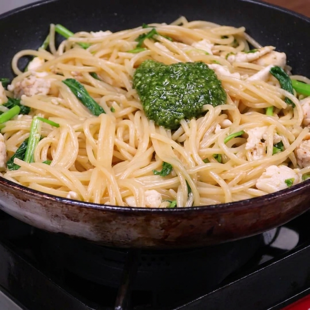Porcja zielonego pesto na wierzchu mieszanki spaghetti, kurczaka i rukoli na patelni.
