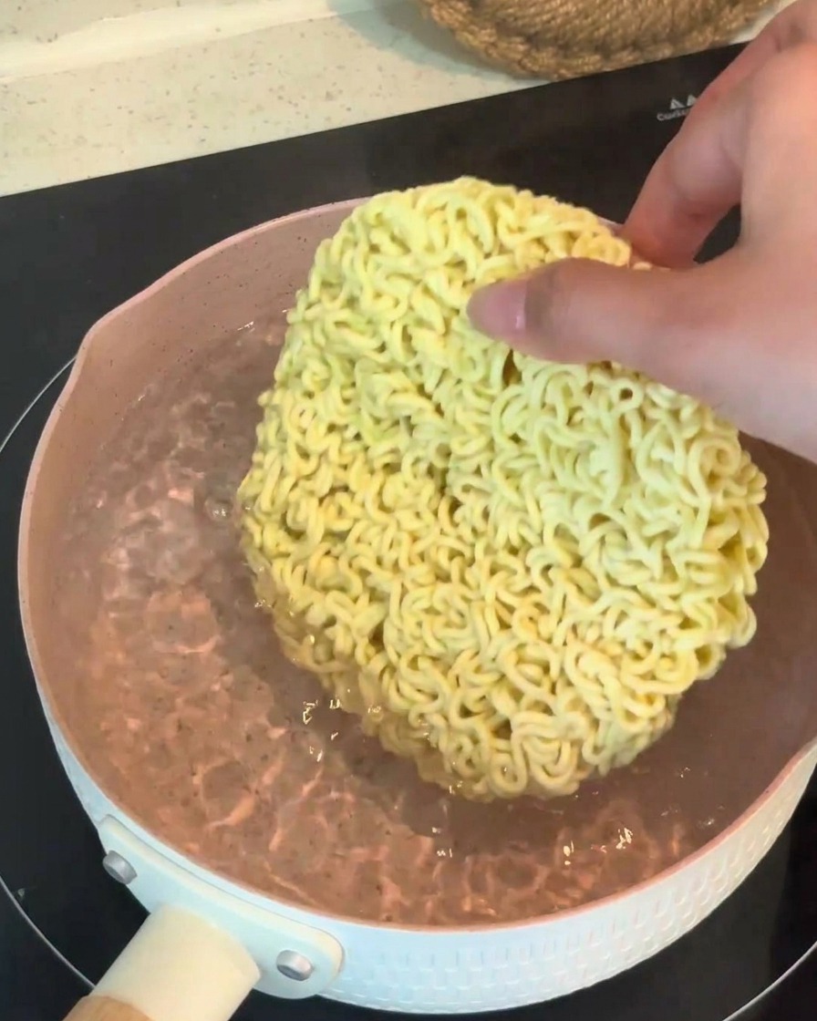 Dłoń wkładająca suchy, okrągły blok makaronu ramen do garnka z wrzącą wodą.