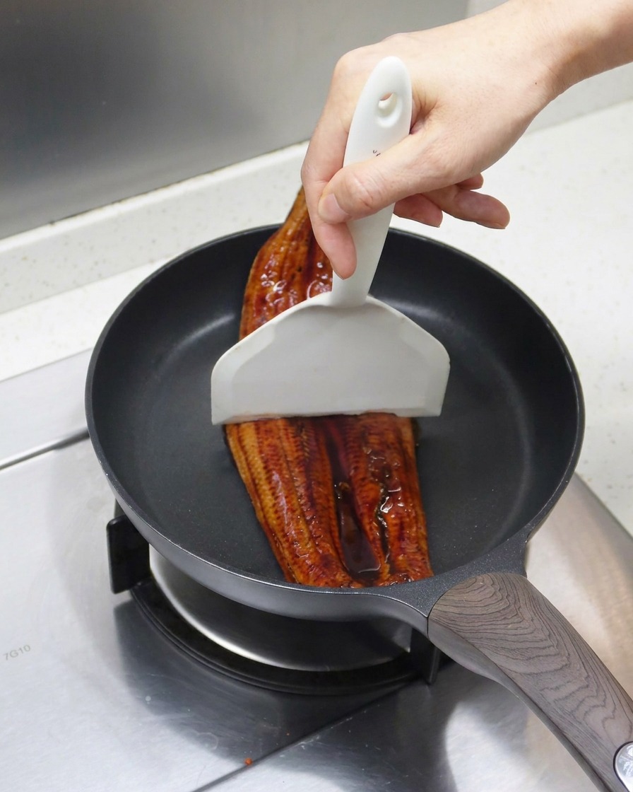 Grillowany węgorz dzielony na dwie części białą łopatką na patelni.