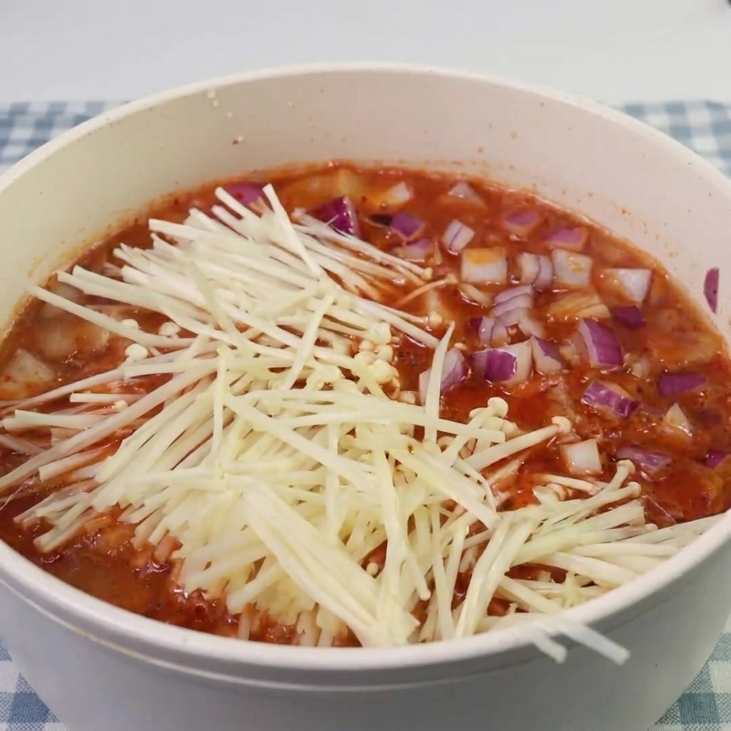 Grzyby enoki i kawałki spamu dodawane do gotującej się zupy kimchi.