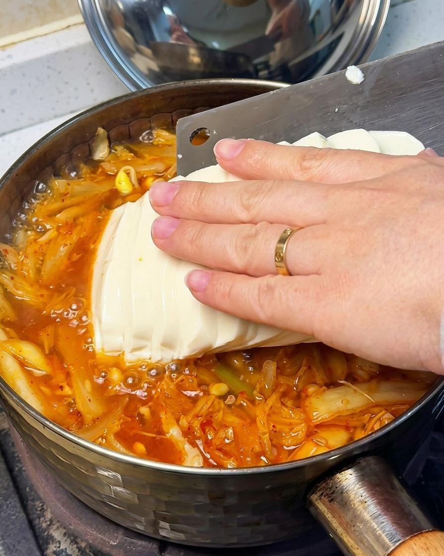 Zbliżenie na dłoń delikatnie układającą plastry jedwabistego tofu do czerwonej zupy kimchi.