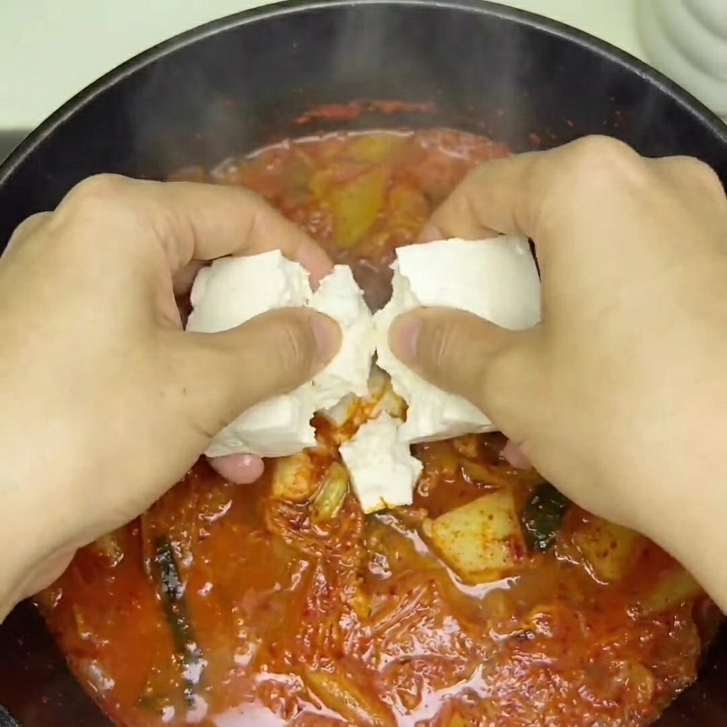 Kostki świeżego tofu wkładane do bulgoczącej, czerwonej zupy kimchi.