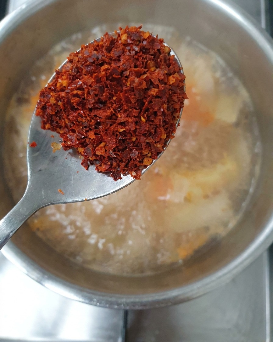 Łyżka z mieszanką gruboziarnistych i drobnych płatków chili trzymana nad zupą.