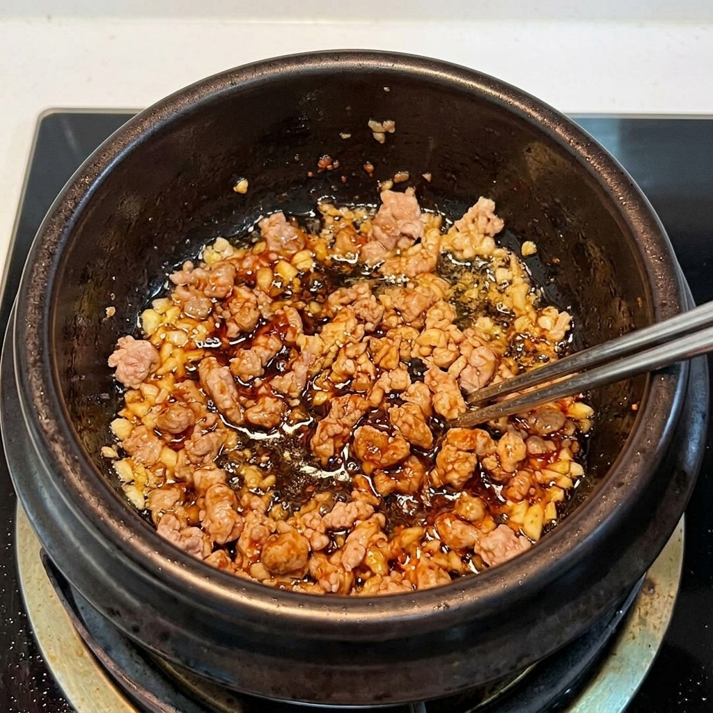 Pokrojone kimchi i mielona wieprzowina mieszane razem w glinianym naczyniu, podsmażane aż do uzyskania aromatu.