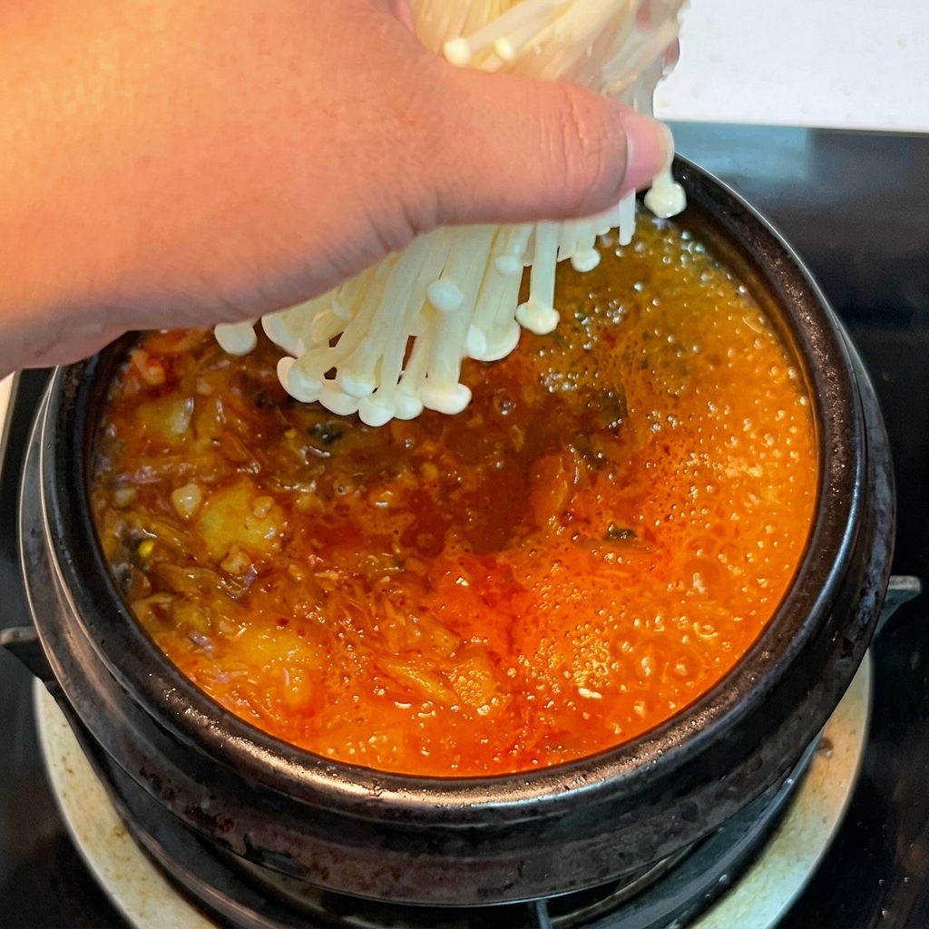 Dłoń wkładająca wiązkę świeżych grzybów enoki do bulgoczącego, czerwonego gulaszu kimchi.