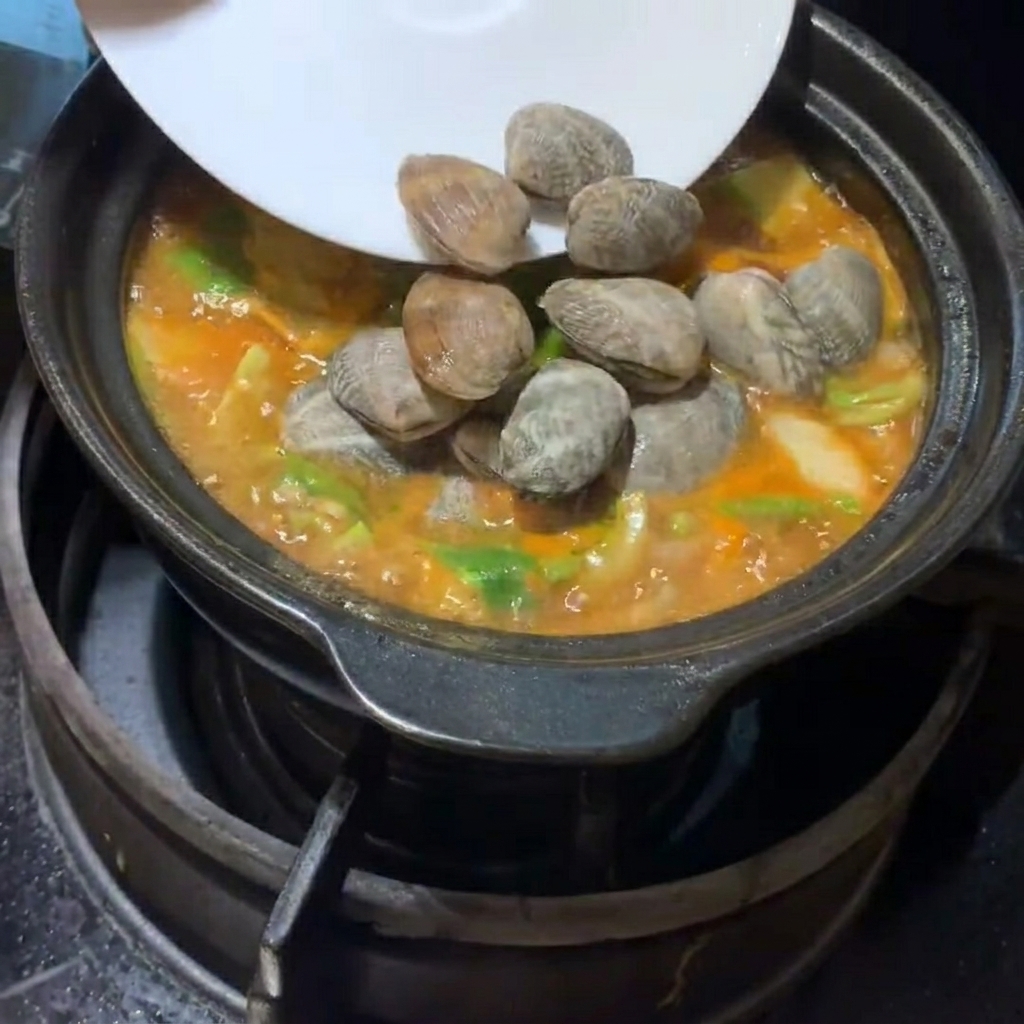 Świeże małże i kostki miękkiego tofu ostrożnie wkładane do bulgoczącego koreańskiego gulaszu.