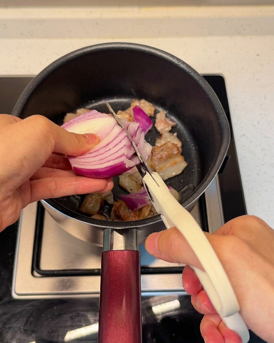 Krojenie świeżej czerwonej cebuli bezpośrednio do garnka z boczkiem przy użyciu nożyczek kuchennych.