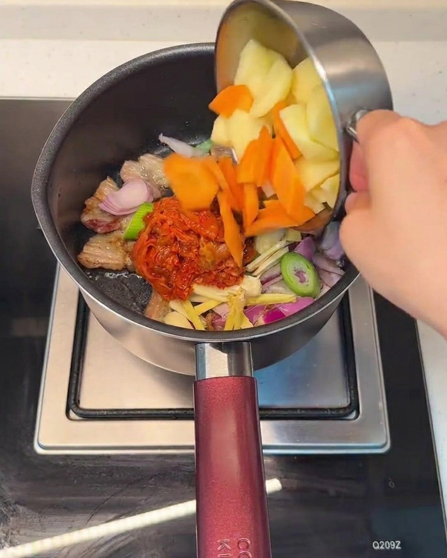 Dodawanie posiekanej marchewki i ziemniaków do garnka z mieszanką boczku i kimchi.