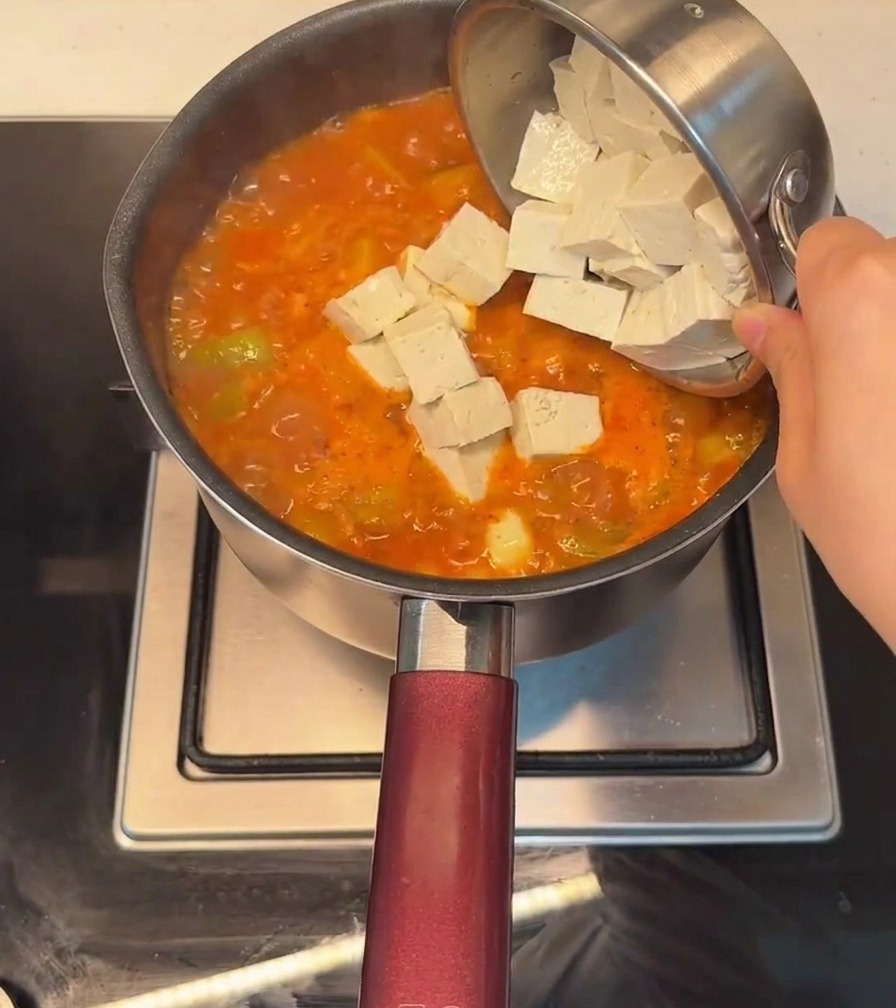 Dodawanie tofu w kostkach do garnka z bulgoczącym, czerwonym, pikantnym gulaszem w koreańskim stylu.