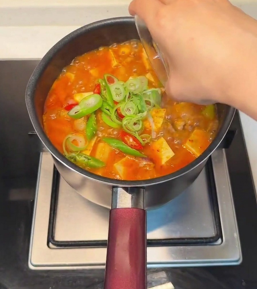 Dłoń posypująca gotowy gulasz z tofu posiekanym szczypiorkiem oraz świeżymi czerwonymi i zielonymi papryczkami chili.