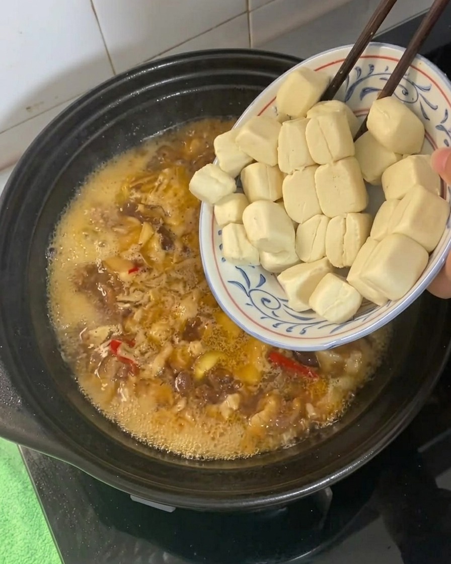 Małe kostki tofu dodawane do bulgoczącego, złocistobrązowego płynu do duszenia w glinianym garnku.