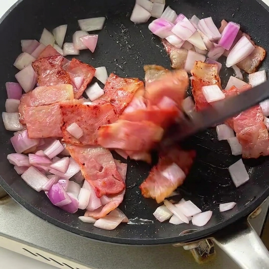Ugotowany makaron fusilli i jasnozielone szparagi dodawane na patelnię z boczkiem i cebulą.