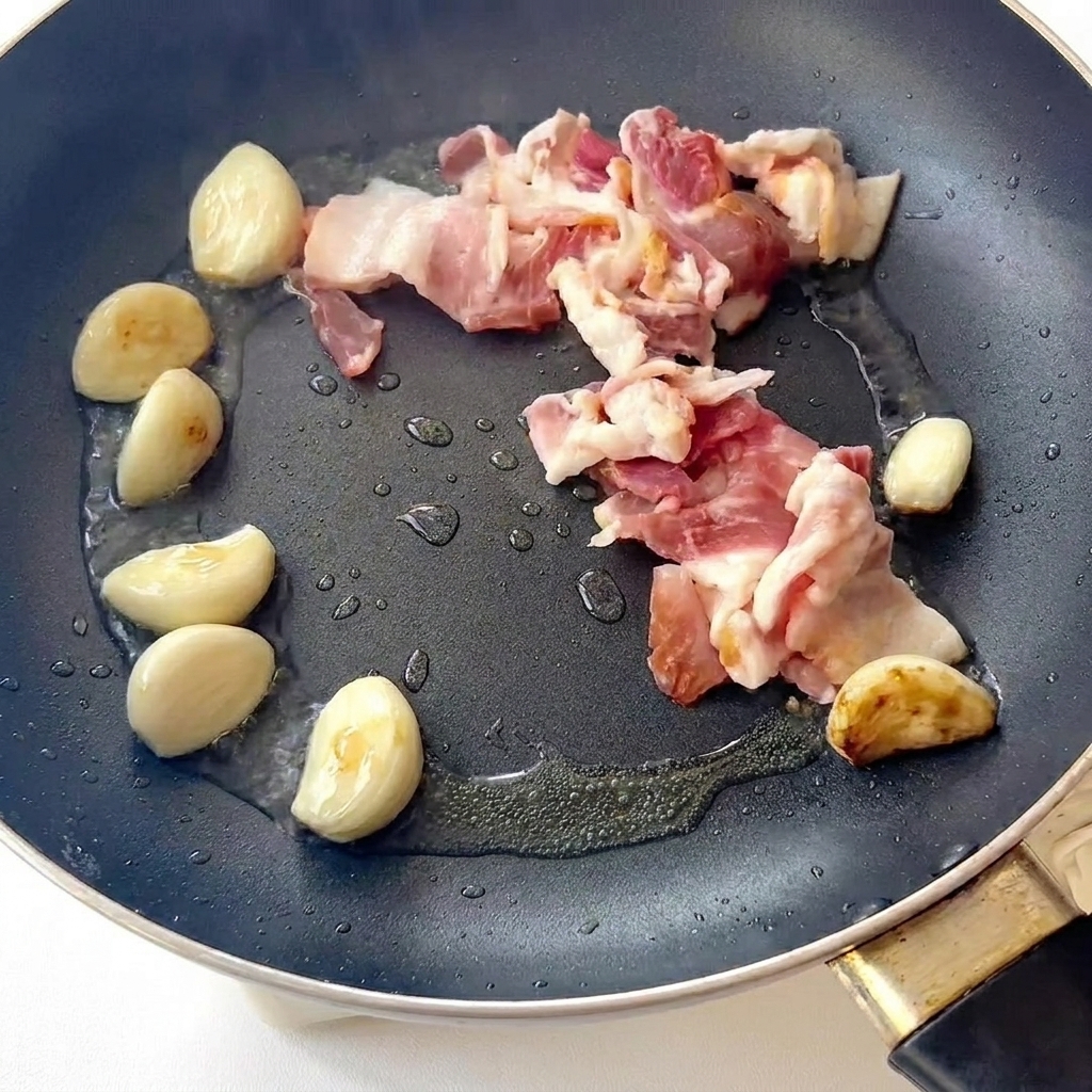 Boczek smażący się na patelni obok podsmażonych ząbków czosnku.