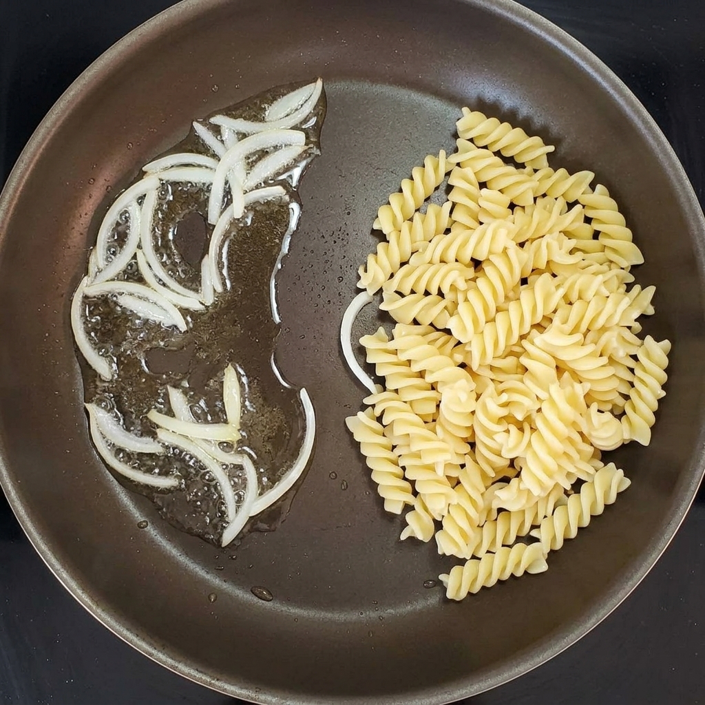 Ugotowany makaron świderki mieszany z cebulą na patelni.