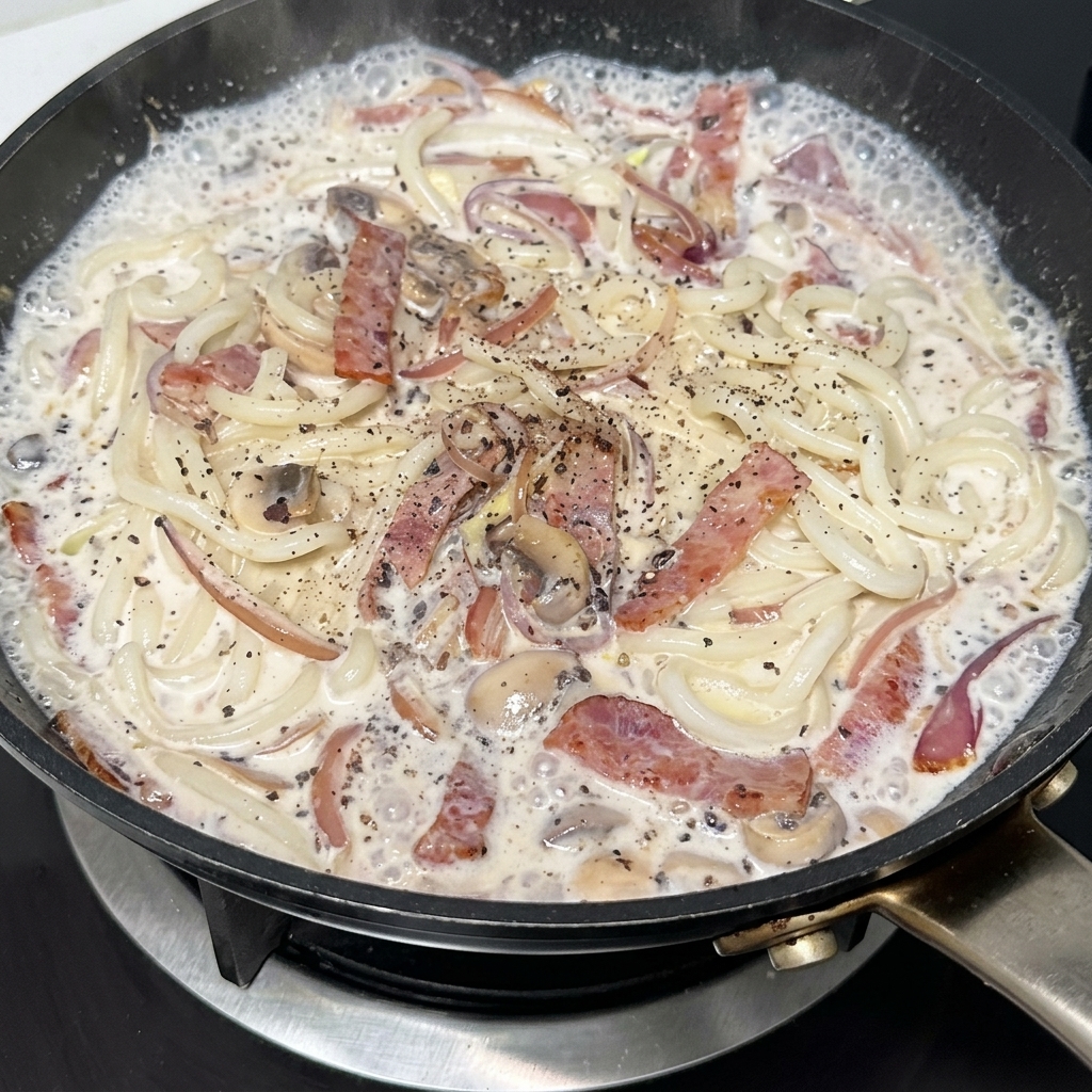 Patelnia kremowego makaronu udon z boczkiem i pieczarkami doprawiana czarnym pieprzem.
