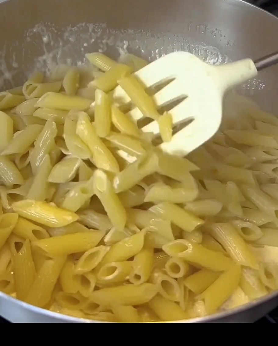 Makaron penne mieszany z kremowym białym sosem i kawałkami boczku na patelni.