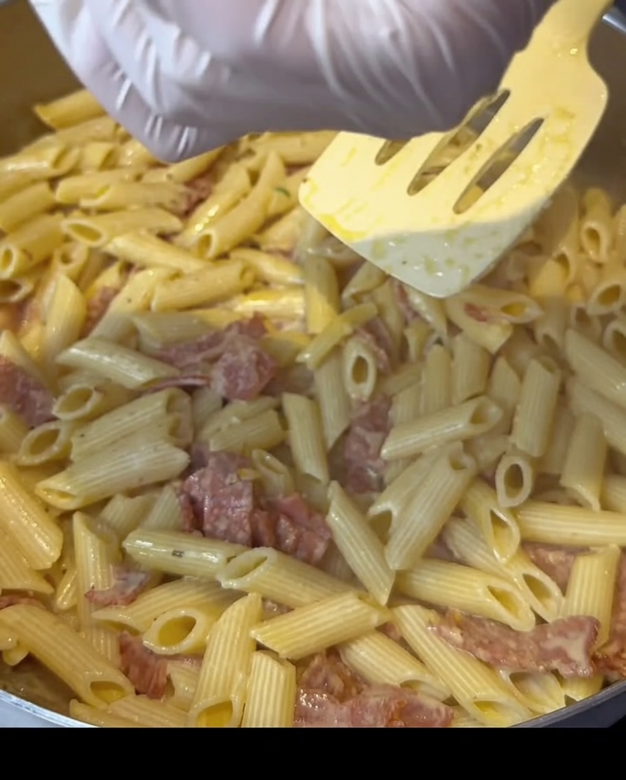 Dłonie w rękawiczkach posypujące przyprawą patelnię z kremowym makaronem i boczkiem.