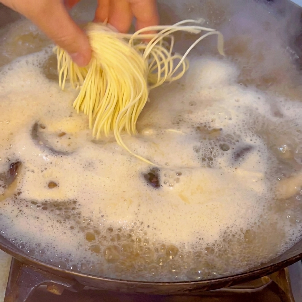 Ręka ostrożnie wrzucająca porcję świeżego makaronu ramen do gotującego się bulionu.