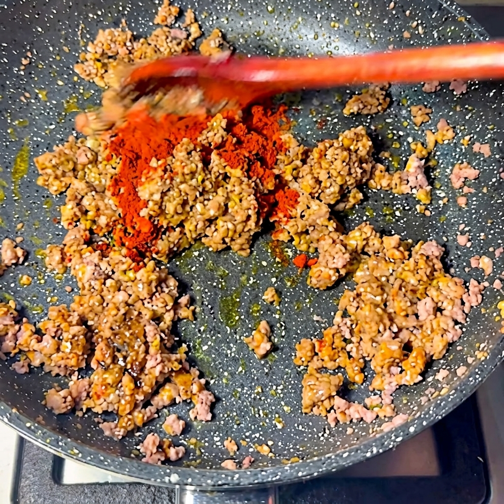 Czerwone chili w proszku posypane na mięsną mieszankę na patelni.