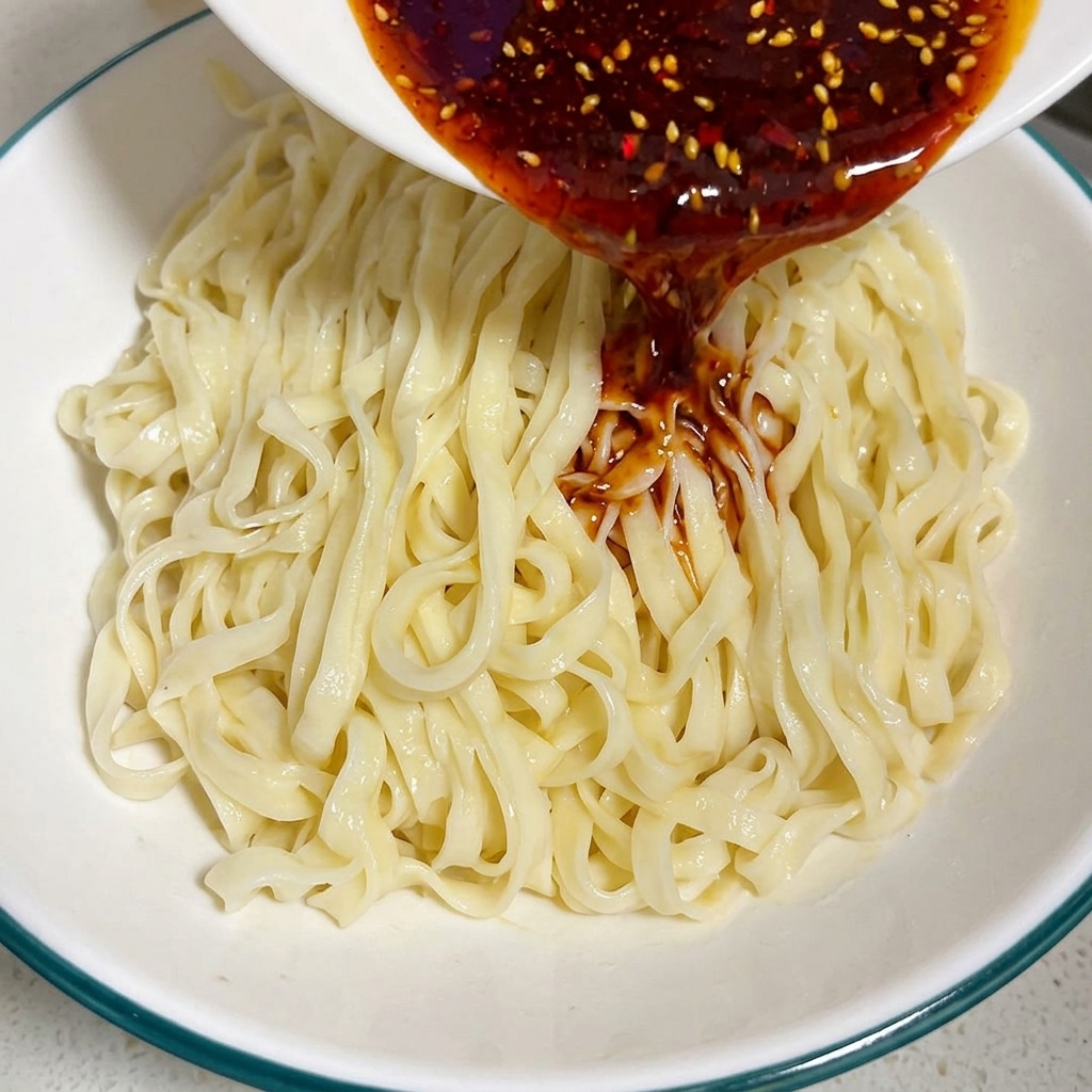 Gęsty, ciemnoczerwony sos curry z chili wylewany z małej miseczki na porcję ugotowanego płaskiego makaronu w większej misce.