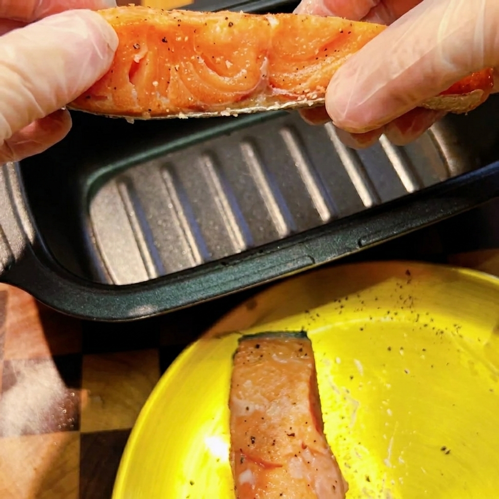 Dłonie w rękawiczkach trzymające przyprawiony surowy filet z łososia nad ciemną, prostokątną patelnią do mikrofalówki.