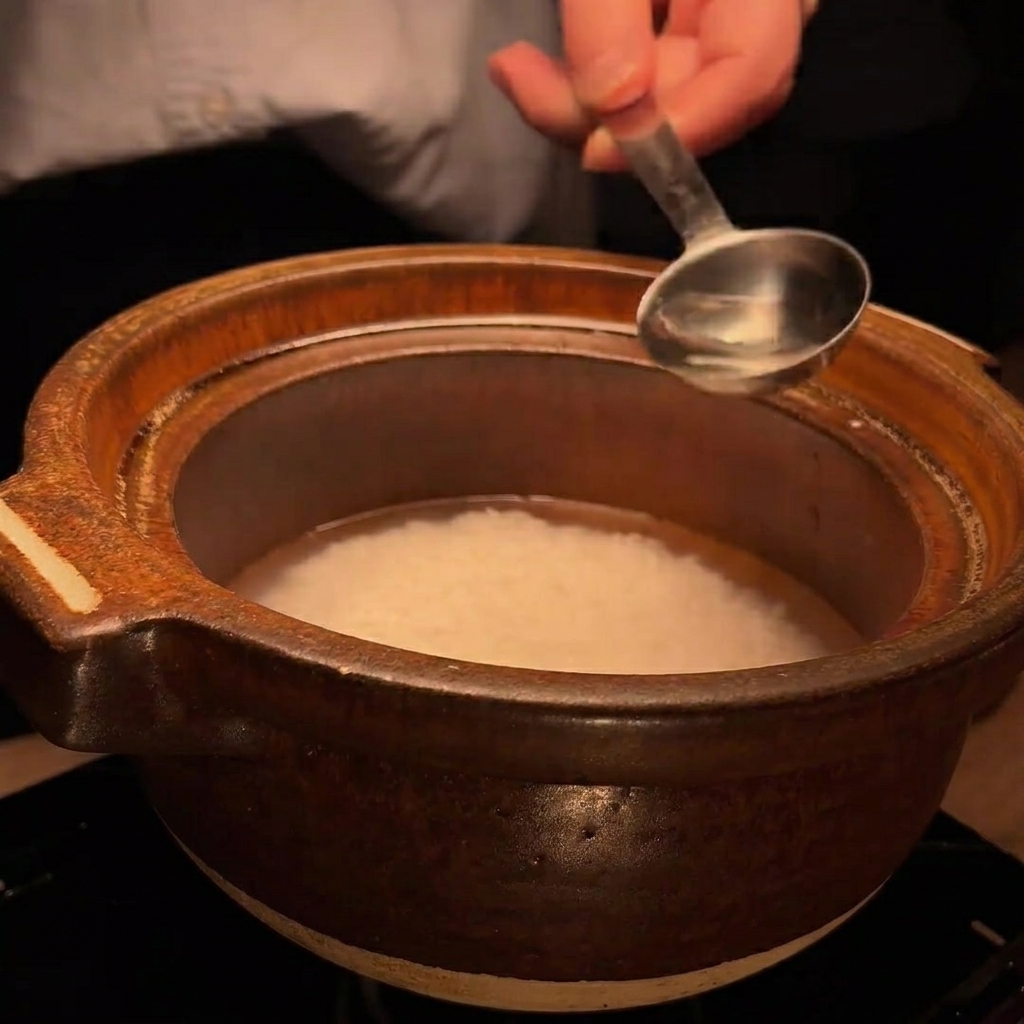 Metalowa łyżka wlewająca niewielką ilość klarownego płynu, sake, do garnka z ryżem.
