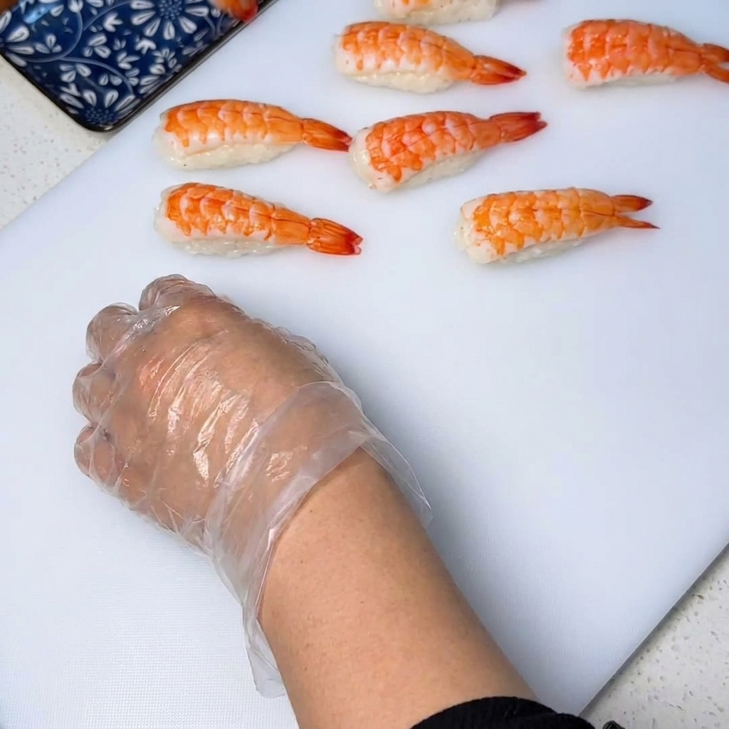 Zbliżenie na gotowaną krewetkę układaną na małej kulce ryżu, dociskaną dłońmi w rękawiczkach, aby nadać sushi odpowiedni kształt.