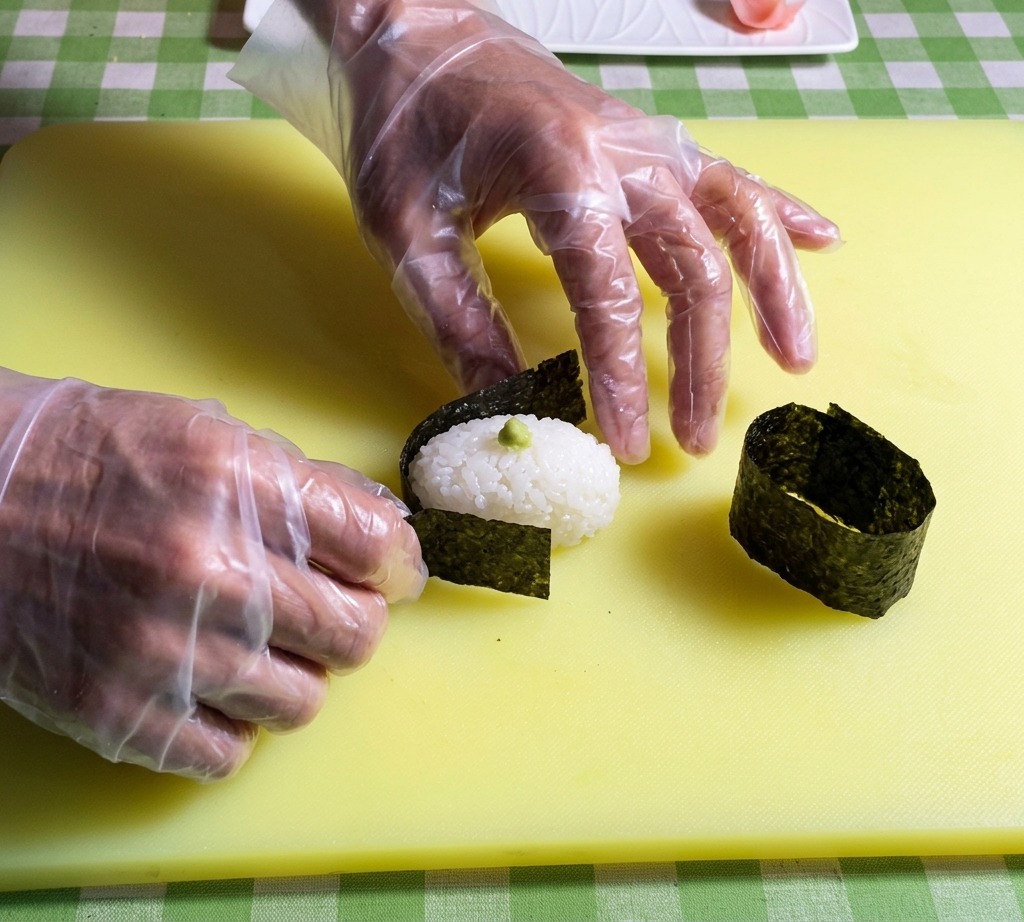 Dłonie owijające pasek ciemnych wodorostów nori wokół krawędzi owalnej bazy z ryżu do sushi.