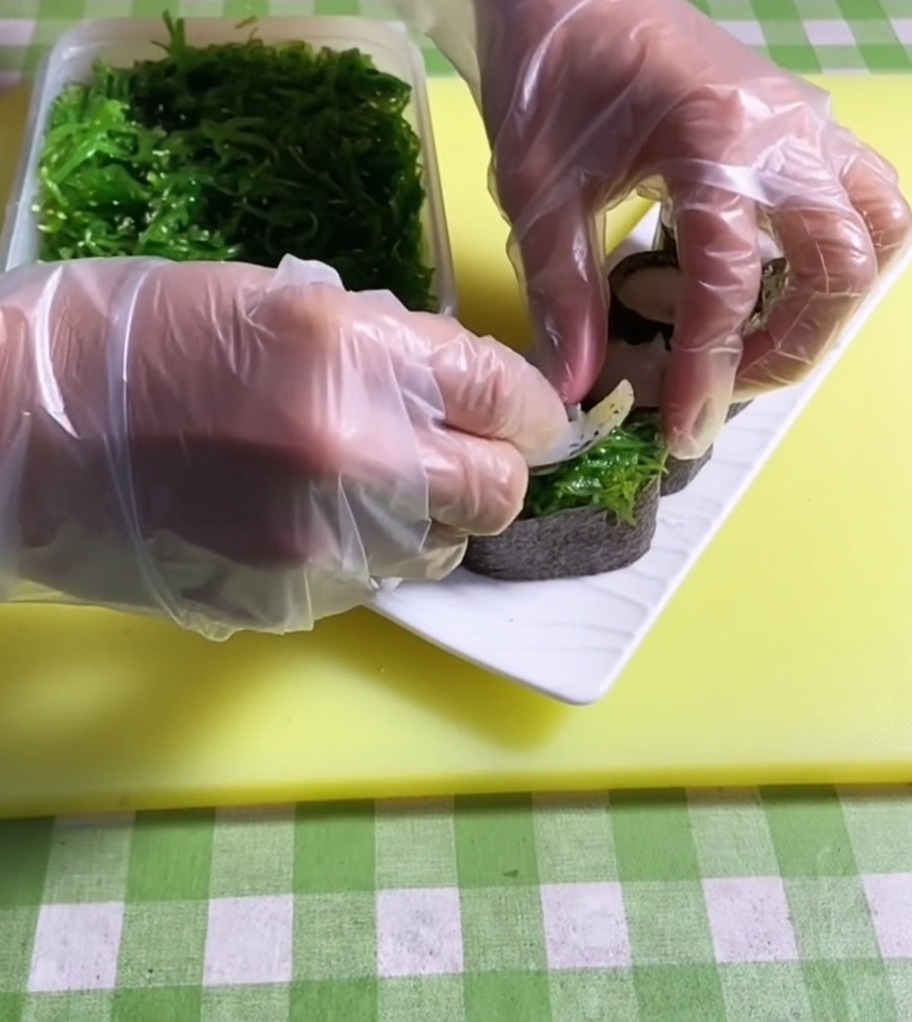 Pęseta nakładająca jaskrawozieloną porcję sałatki z wodorostów na wierzch bazy do sushi.