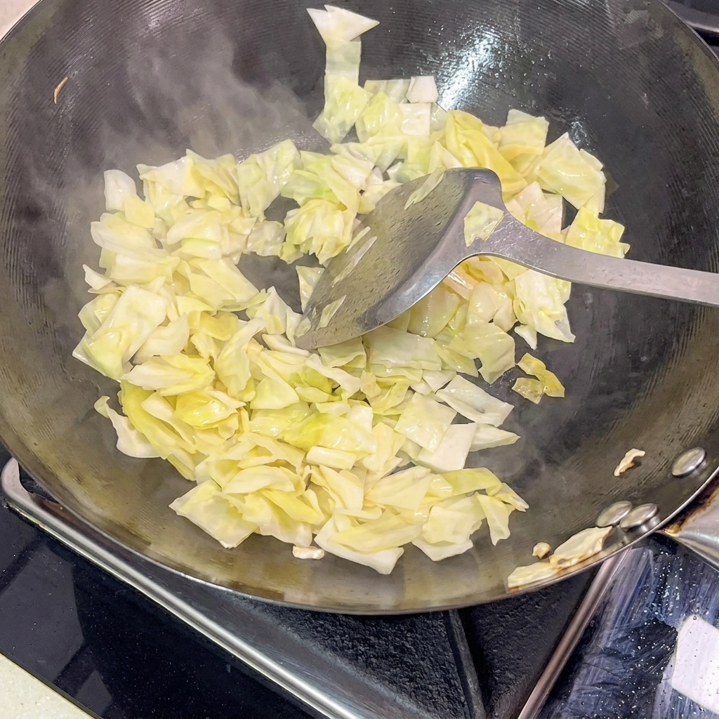 Metalowa łopatka mieszająca jasną kiszoną kapustę w gorącym czarnym woku, z widoczną parą unoszącą się z patelni.