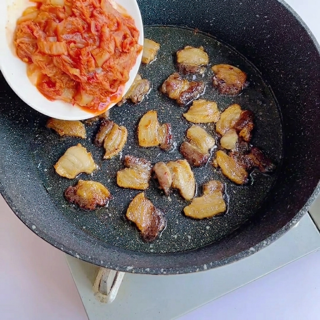 Posiekane kimchi dodawane na patelnię z zrumienionymi plastrami boczku.