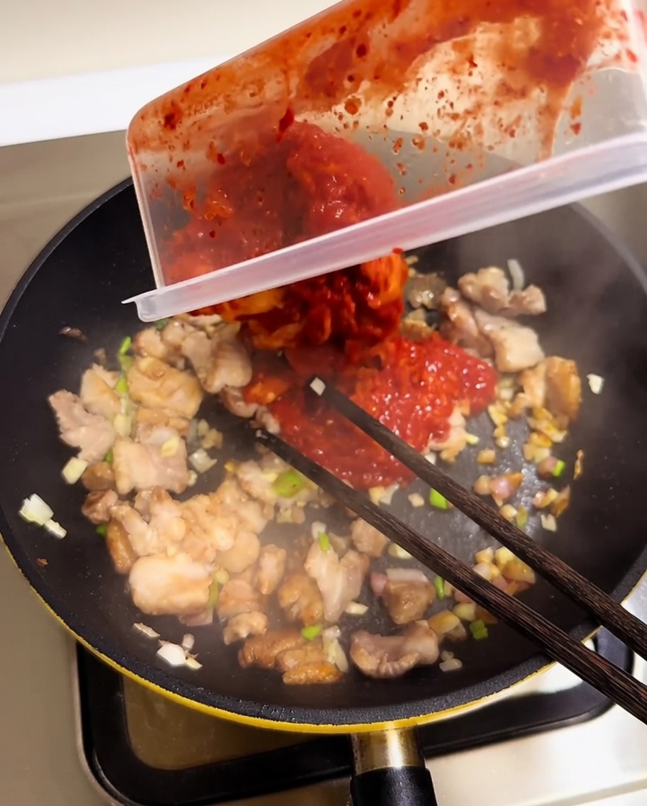 Posiekane czerwone kimchi dodawane do patelni z boczkiem i warzywami.