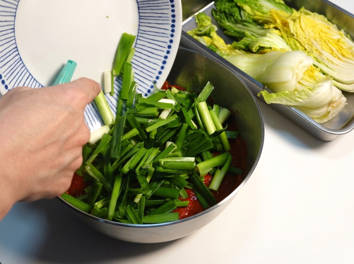 Posiekany zielony szczypiorek dodawany do gęstej czerwonej pasty kimchi w metalowej misce.